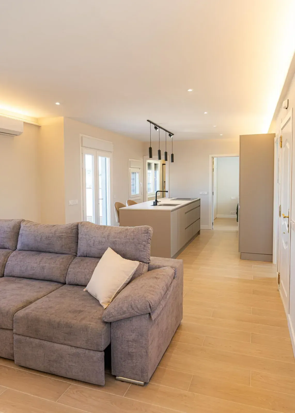 Cocina y sala de estar amplias y luminosas, con sofá gris, suelos de madera clara y lámparas colgantes sobre la isla
