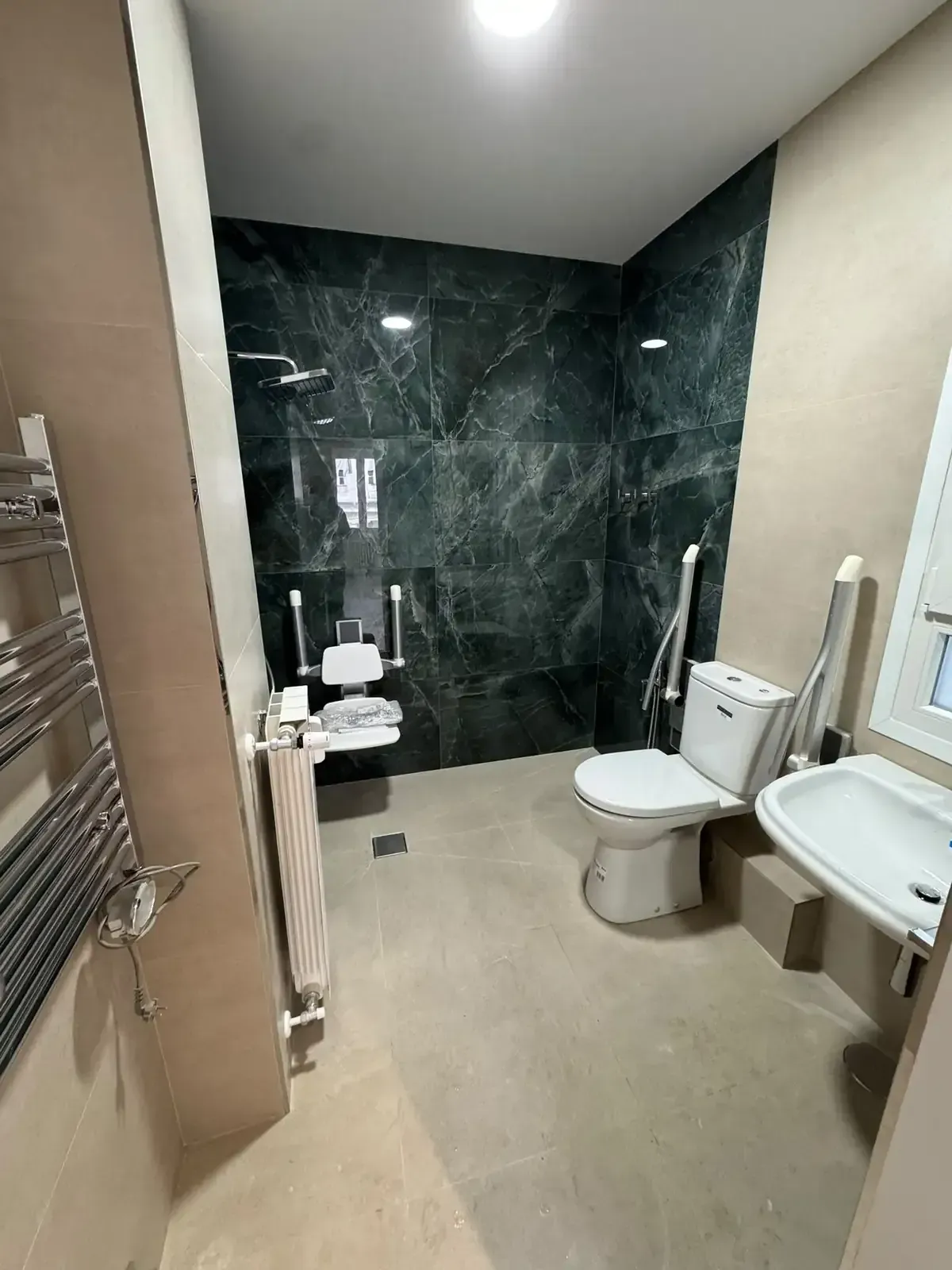 Baño moderno con suelo beige, pared de ducha con azulejos verde oscuro, inodoro, lavabo, radiador toallero y barras de apoyo