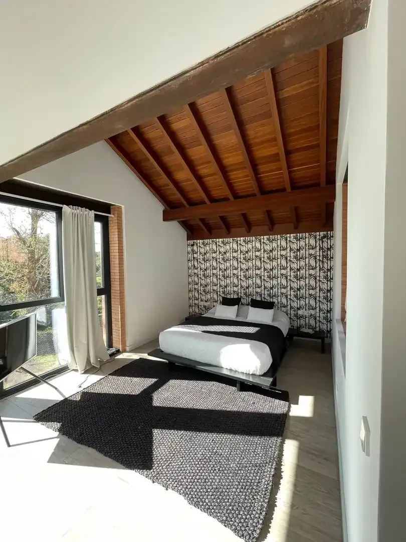 Luminoso dormitorio tipo loft con techo de madera inclinado, puerta abierta y cama junto a una cortina de pared estampada.