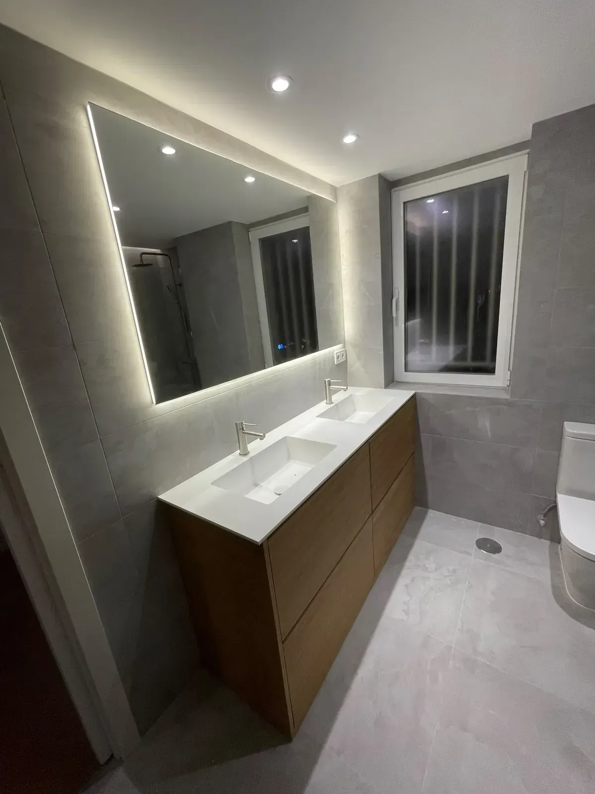 Mueble de baño moderno con un gran espejo retroiluminado, lavabo blanco, armarios de madera y paredes revestidas