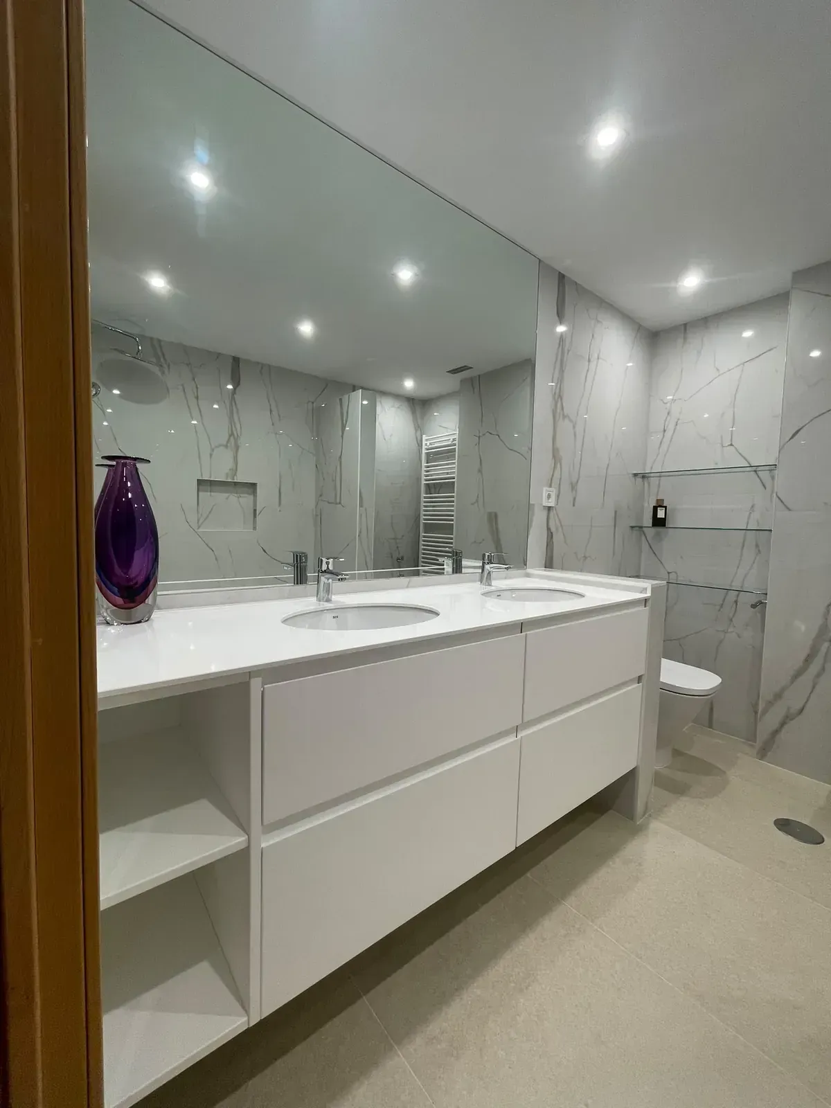 Baño moderno con lavabo doble, espejo grande, azulejos de mármol blanco y ducha a ras de suelo