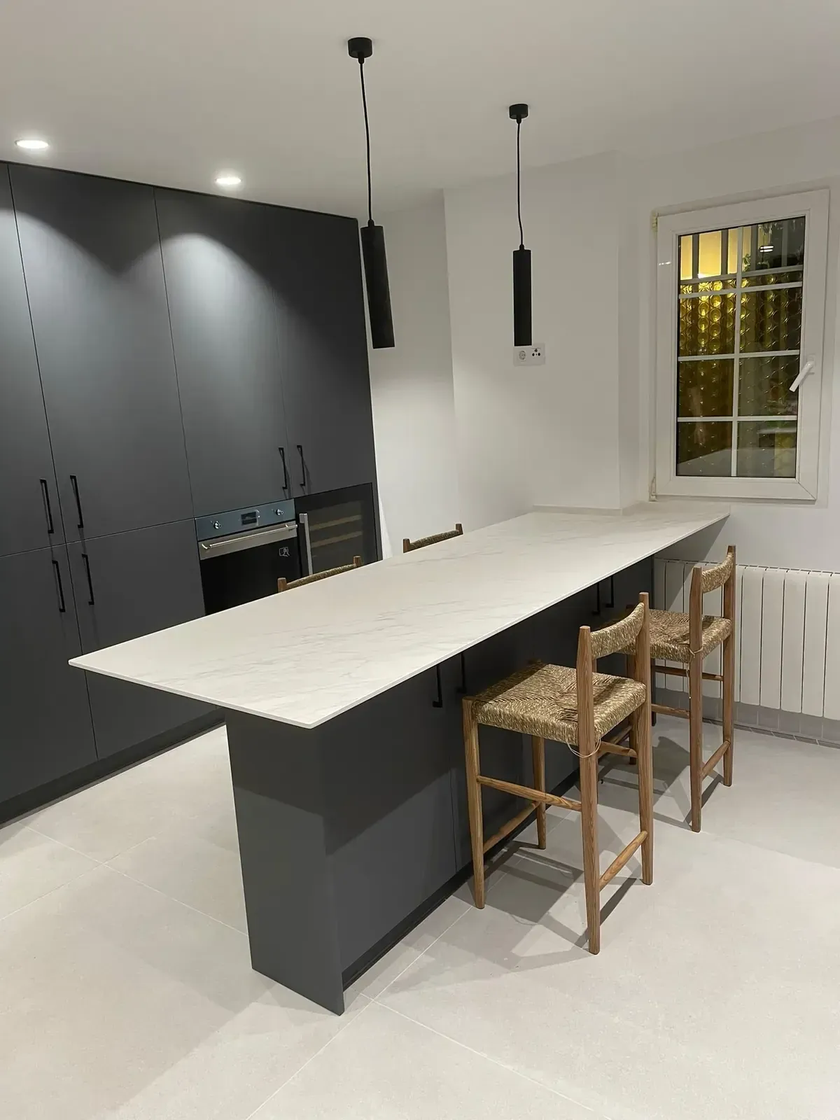 Cocina moderna con una gran isla blanca, armarios oscuros, lámparas colgantes y tres taburetes de mimbre.