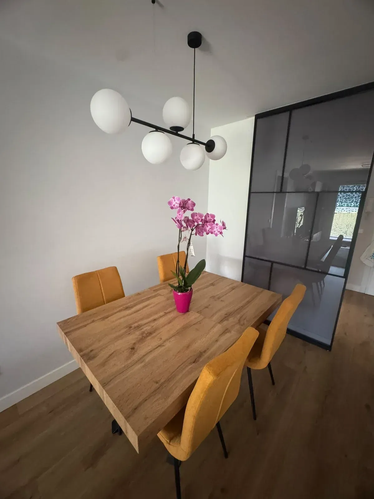 Comedor moderno con mesa de madera, sillas color mostaza, lámpara de araña negra y orquídeas rosas sobre la mesa
