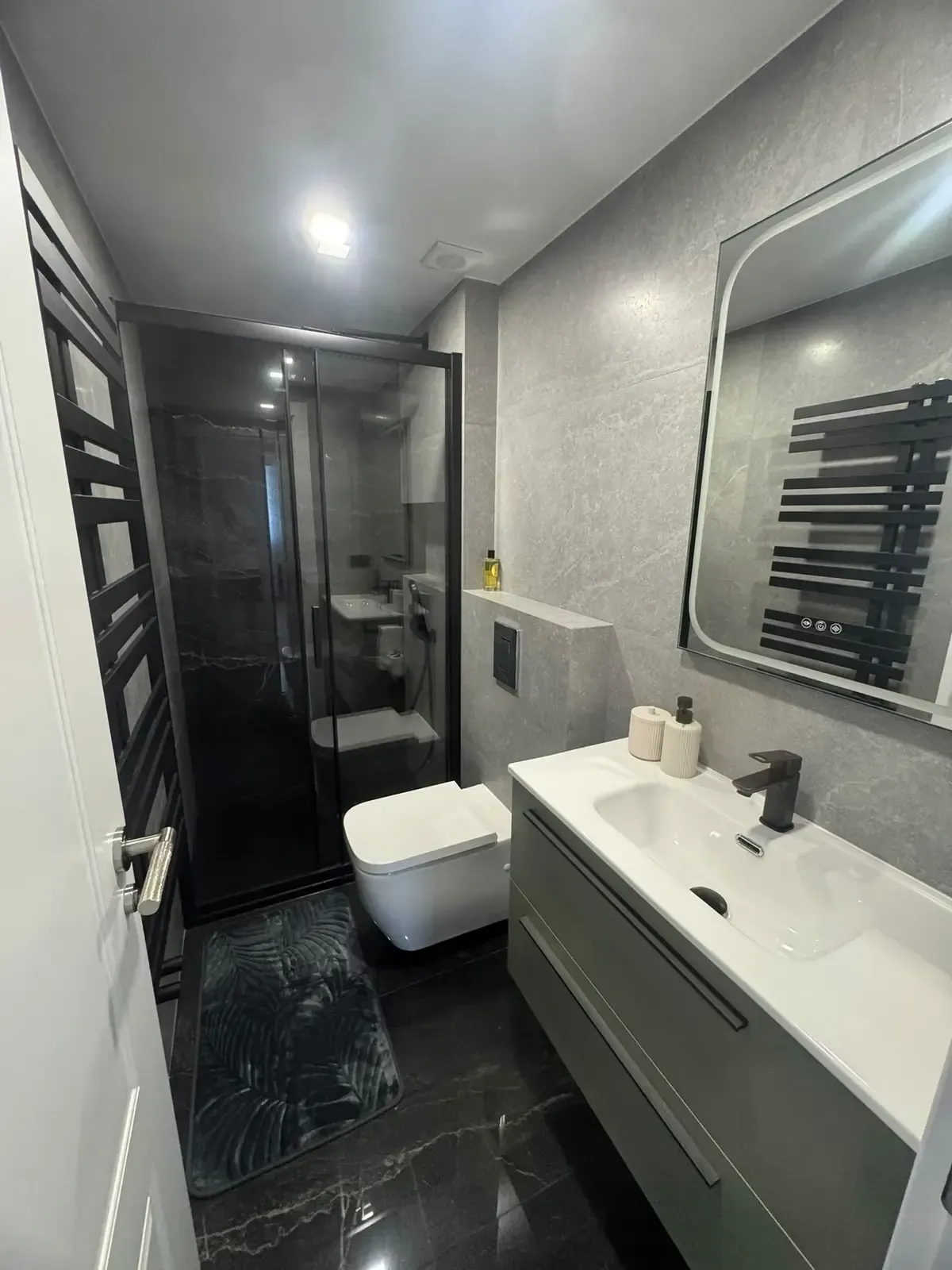 Baño moderno y estrecho con ducha negra, espejo de pared, lavabo blanco e inodoro sobre suelo oscuro.