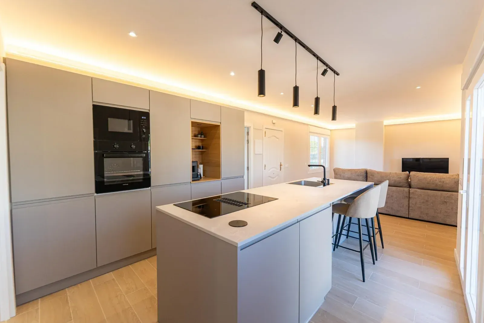 Cocina moderna blanca con isla, lámparas colgantes, hornos empotrados y una zona de estar con un sofá al fondo