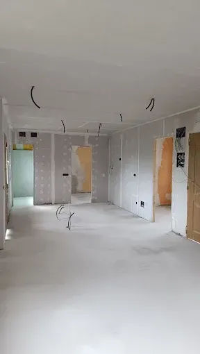 Habitación vacía sin terminar, con paredes y suelo blancos, cables a la vista en el techo y puertas abiertas.