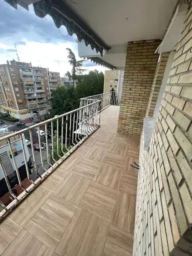 Balcón con barandilla blanca, pared de ladrillo y suelo de baldosas, con vistas a edificios de apartamentos y árboles.