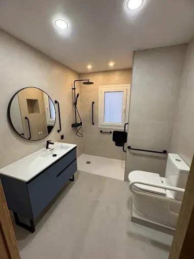 Baño moderno con lavabo con mueble, espejo redondo, ducha a ras de suelo e inodoro, iluminado por luces brillantes en el techo