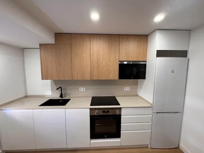 Cocina moderna y minimalista con armarios blancos, armarios superiores de madera, encimera negra y electrodomésticos de acero inoxidable