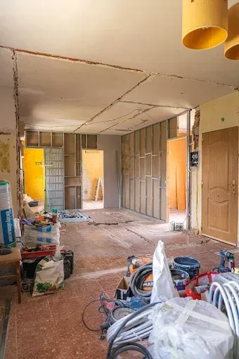 Habitación interior sin terminar, con la estructura a la vista, placas de yeso, materiales de construcción y una puerta que da a otra habitación.