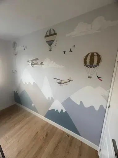 Mural infantil con montañas, nubes, globos aerostáticos y aviones en una pared gris