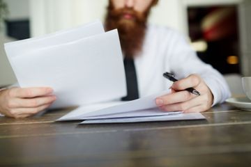 Hombre con barba roja, revisando documentos en una mesa, sosteniendo un bolígrafo.