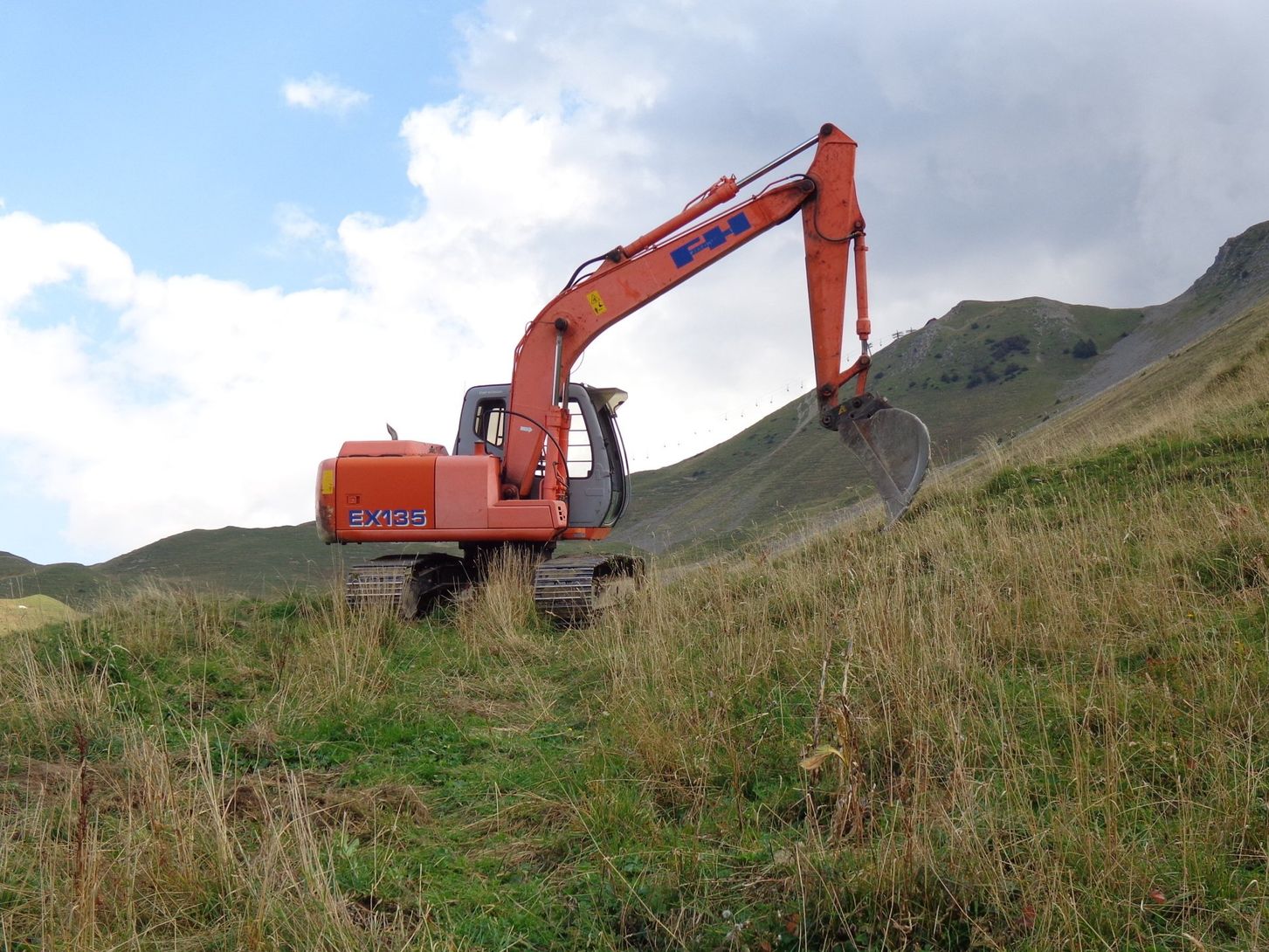 Une pelleteuse orange creuse dans les hautes herbes à flanc de colline sous un ciel nuageux.