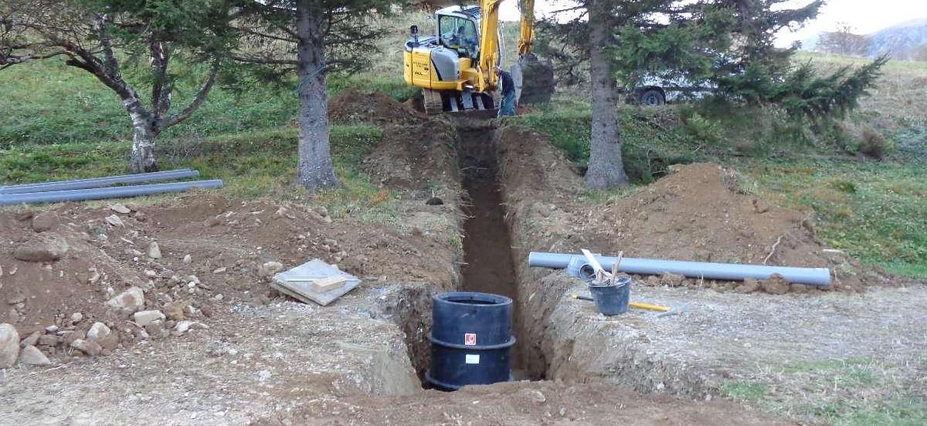 Une pelleteuse jaune creuse une tranchée entre des arbres au bord d'un chemin de terre, avec des tuyaux et un godet noir à l'avant.