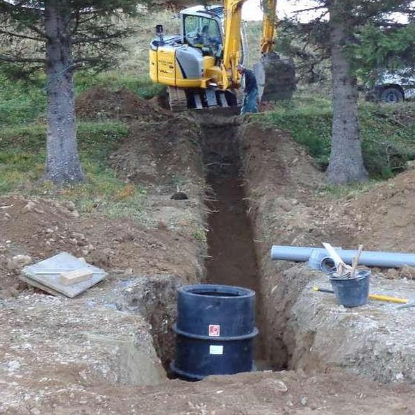 Une excavatrice creuse une tranchée, un fût noir se trouvant au premier plan, près des arbres.