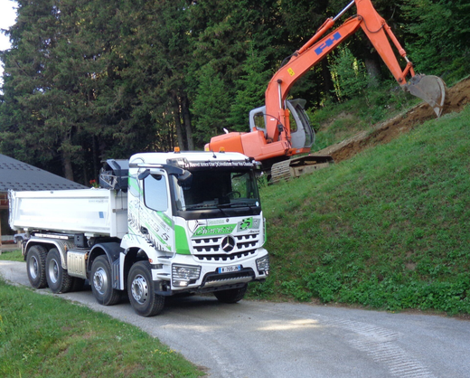 Une pelleteuse orange déverse de la terre dans un camion-benne blanc sur un talus herbeux en bord de route.