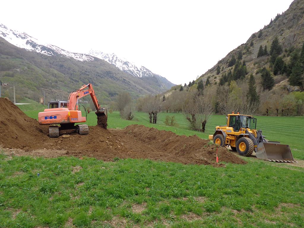 Une excavatrice orange et une chargeuse jaune déplacent de la terre sur un chantier de construction en montagne.