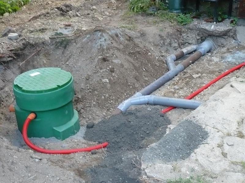 Tranchée de chantier avec boîtier électrique vert et tuyaux gris apparents le long d'un trottoir.
