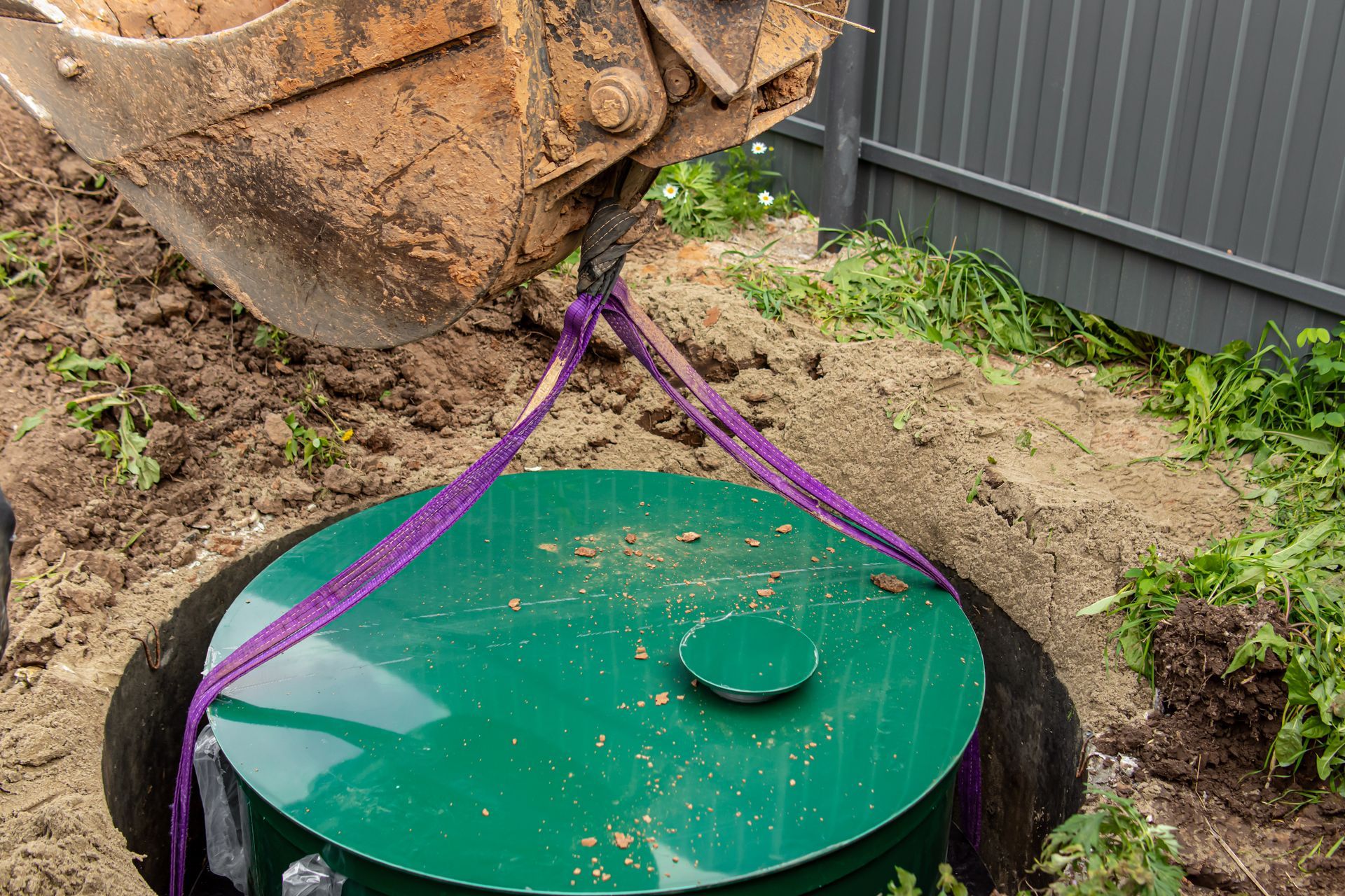 Une excavatrice soulève un réservoir cylindrique vert d'un trou creusé dans une cour boueuse à l'aide de sangles violettes.