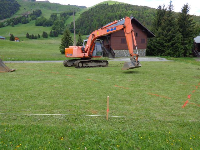 Une pelleteuse orange sur un champ herbeux, avec une maison et des arbres en arrière-plan.