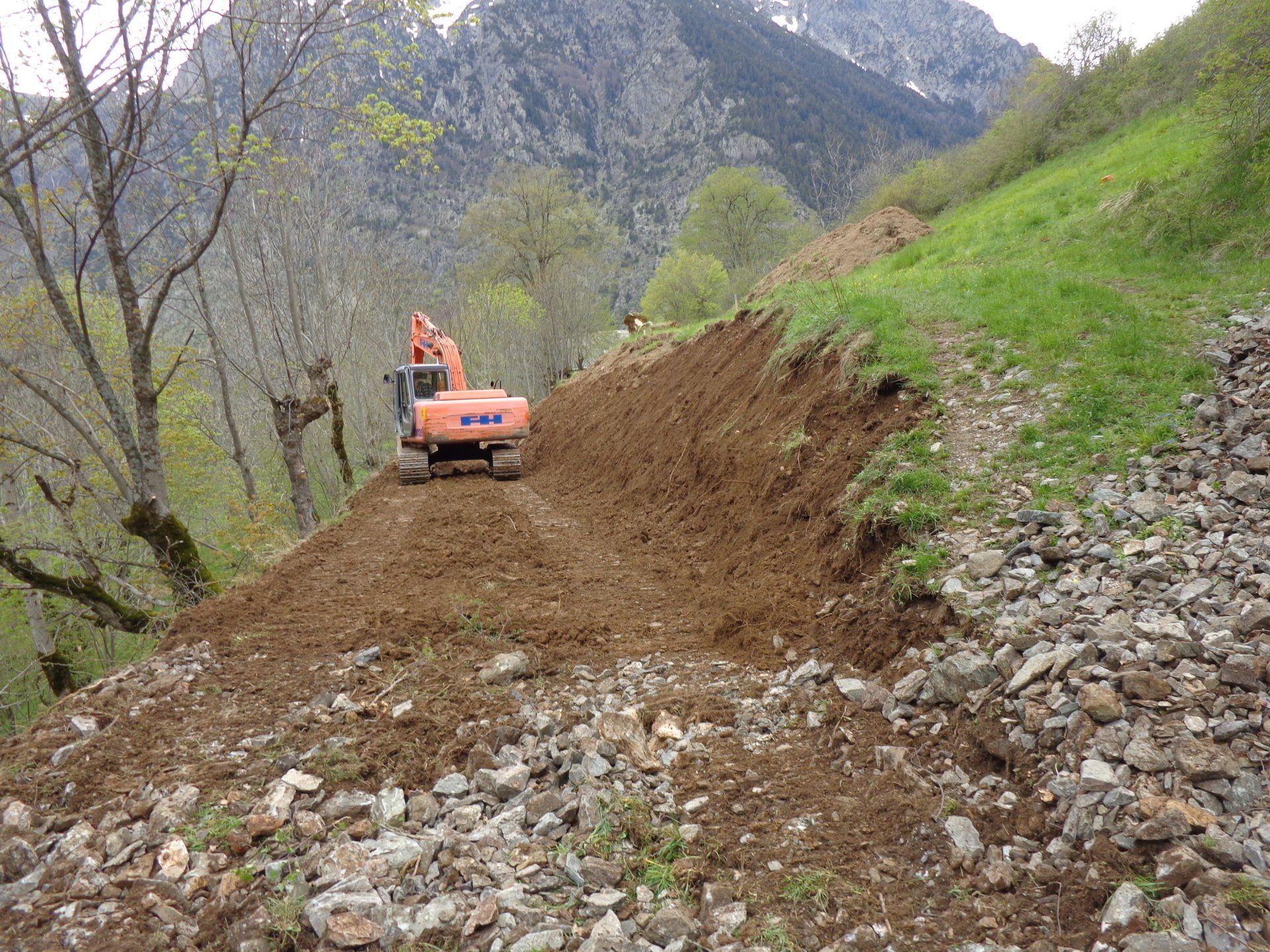 Une pelleteuse orange déblayait un chemin de terre à flanc de colline rocheuse dans une vallée de montagne.