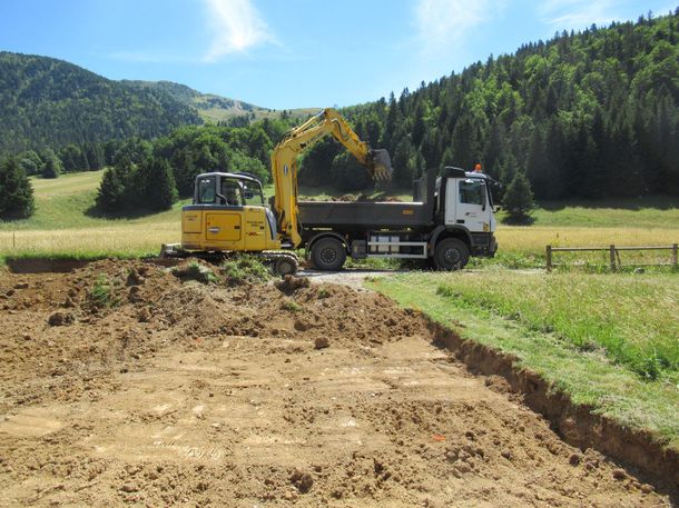 Une pelleteuse jaune charge de la terre dans un camion-benne sur un chemin de terre, au milieu d'un champ verdoyant avec des collines en arrière-plan.