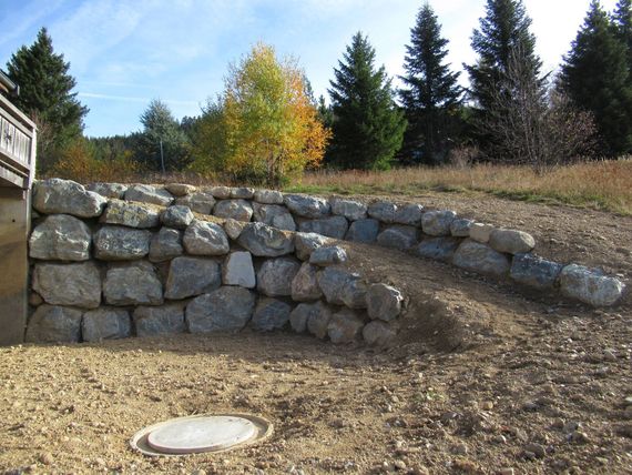 Mur de soutènement incurvé en pierre, bordant une zone de gravier et un versant herbeux avec des arbres en arrière-plan.