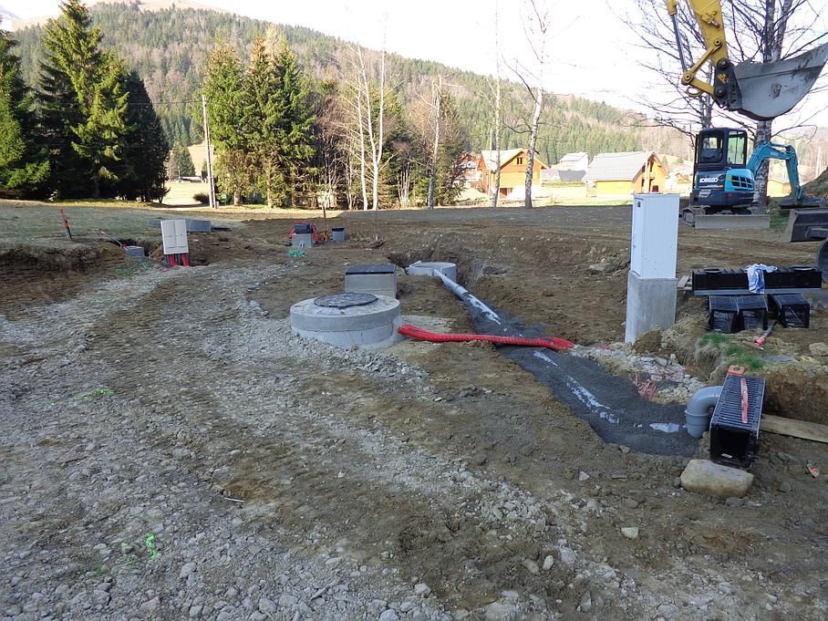 Chantier de construction avec canalisations et fondations en béton sur un terrain en terre battue près d'arbres et de collines.