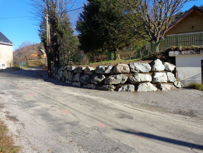 Route de campagne longeant un mur de soutènement en pierre, des arbres et des maisons par une journée ensoleillée
