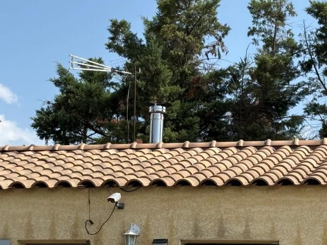 Toit de tuiles en terre cuite avec une cheminée en métal se détachant sur les arbres et le ciel bleu.
