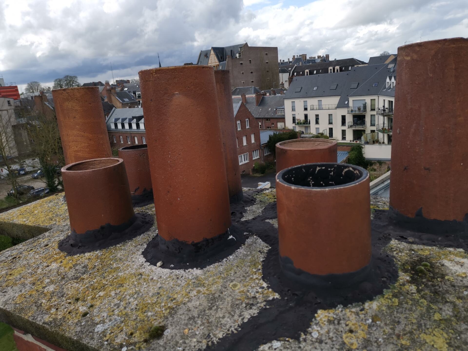 Plusieurs cheminées en briques sur un toit, avec un arrière-plan de ville sous un ciel nuageux.