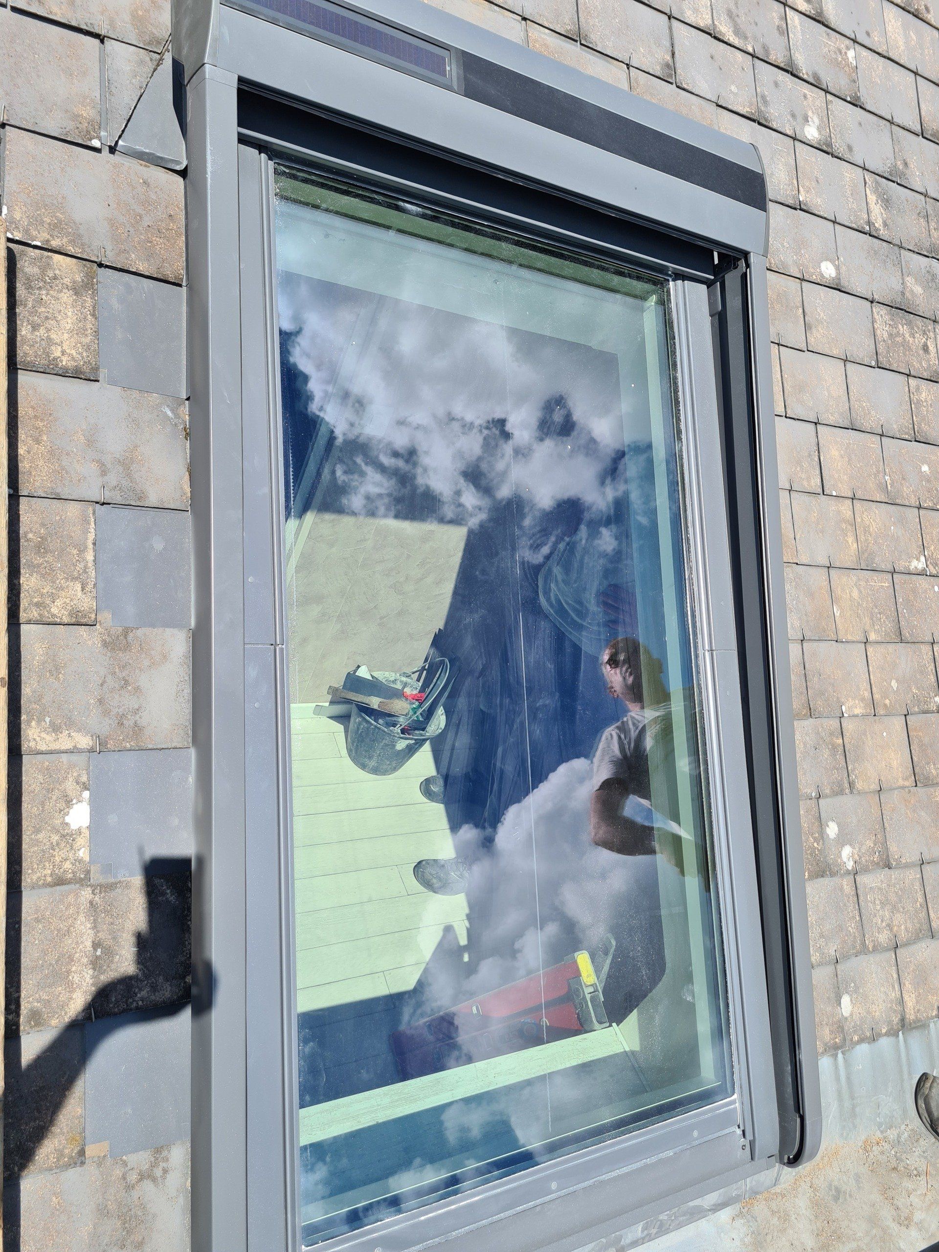 Fenêtre avec store enrouleur gris partiellement abaissé reflétant une personne, un ciel bleu et de l'herbe verte.