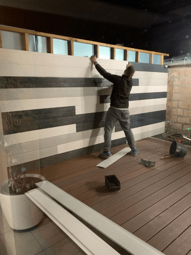 Un homme installe des panneaux imitation bois sur un mur ; le décor semble être une terrasse extérieure.