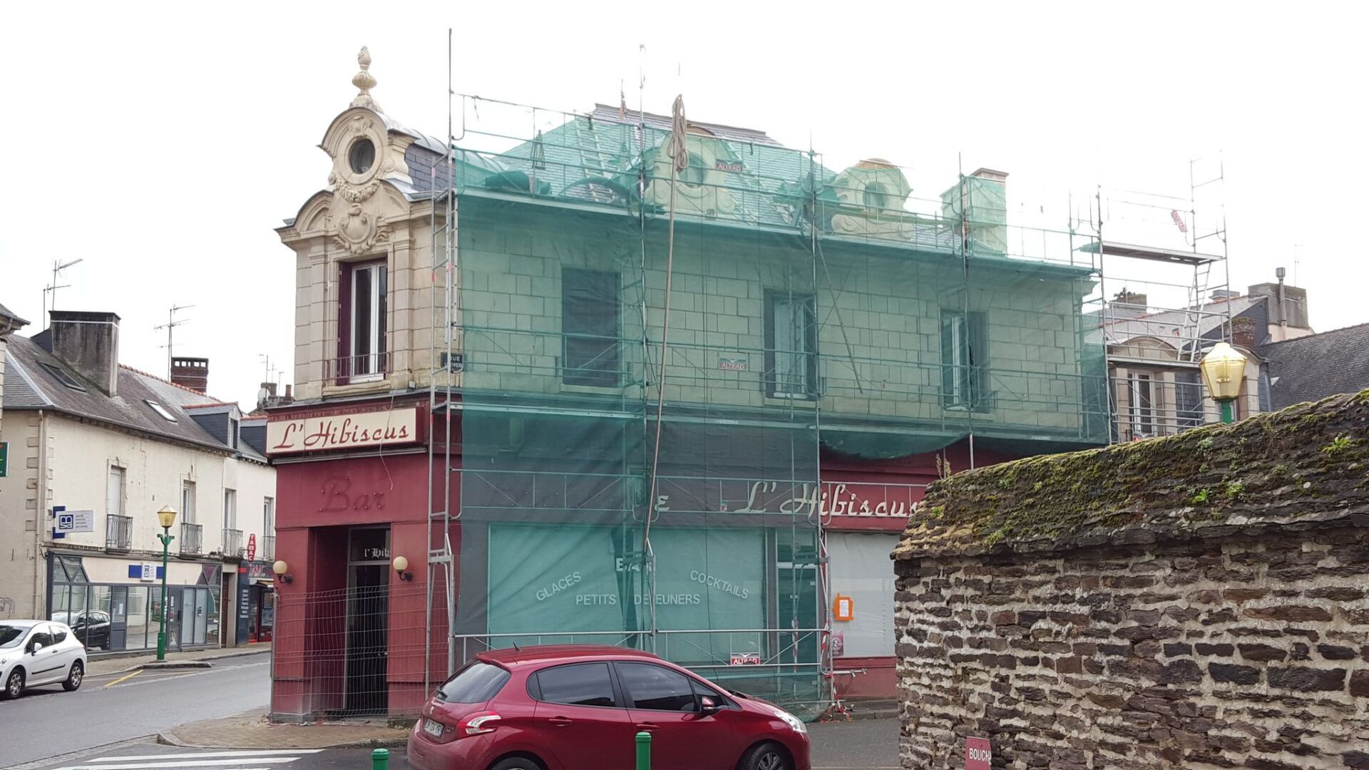 Bâtiment en rénovation, recouvert d'échafaudages et de filets verts, au coin d'une rue, avec des voitures garées et des commerces visibles.