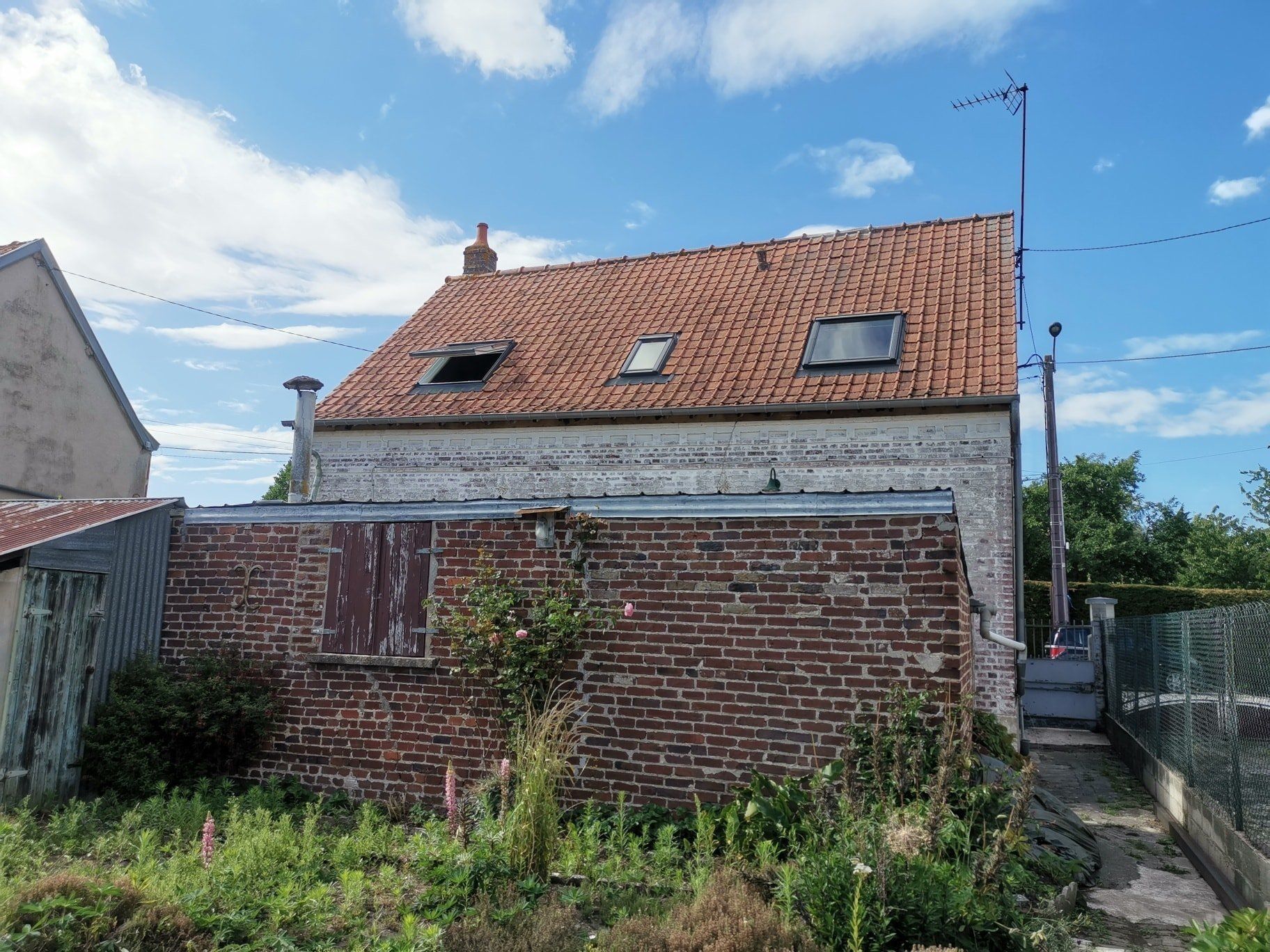 Maison en briques avec toit en tuiles rouges, petit hangar et cour envahie par la végétation sous un ciel bleu.
