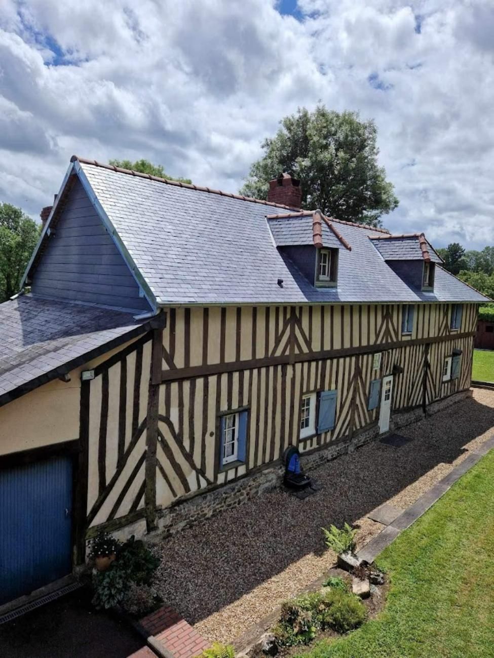 Maison à colombages avec toit en ardoise et volets bleus, située dans une cour herbeuse sous un ciel nuageux.