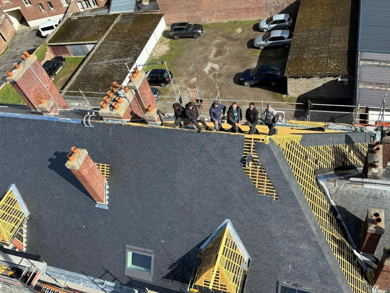 Des ouvriers sur un toit avec du bois exposé, des cheminées et des voitures garées en dessous.
