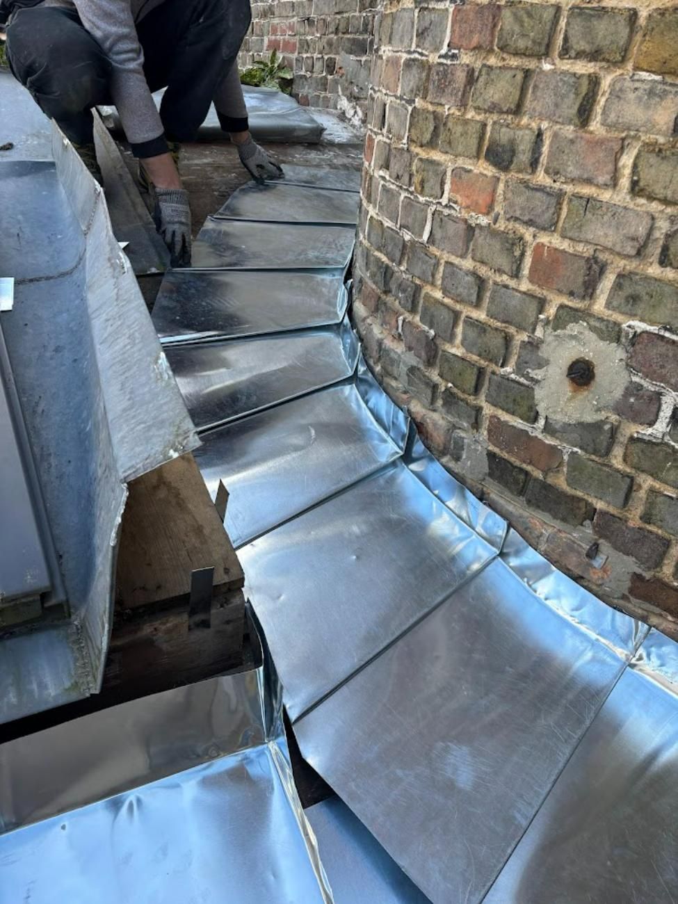 Couvreur installant des solins en plomb le long d'un mur de briques. Plomb argenté, briques brunes et gants gris visibles.