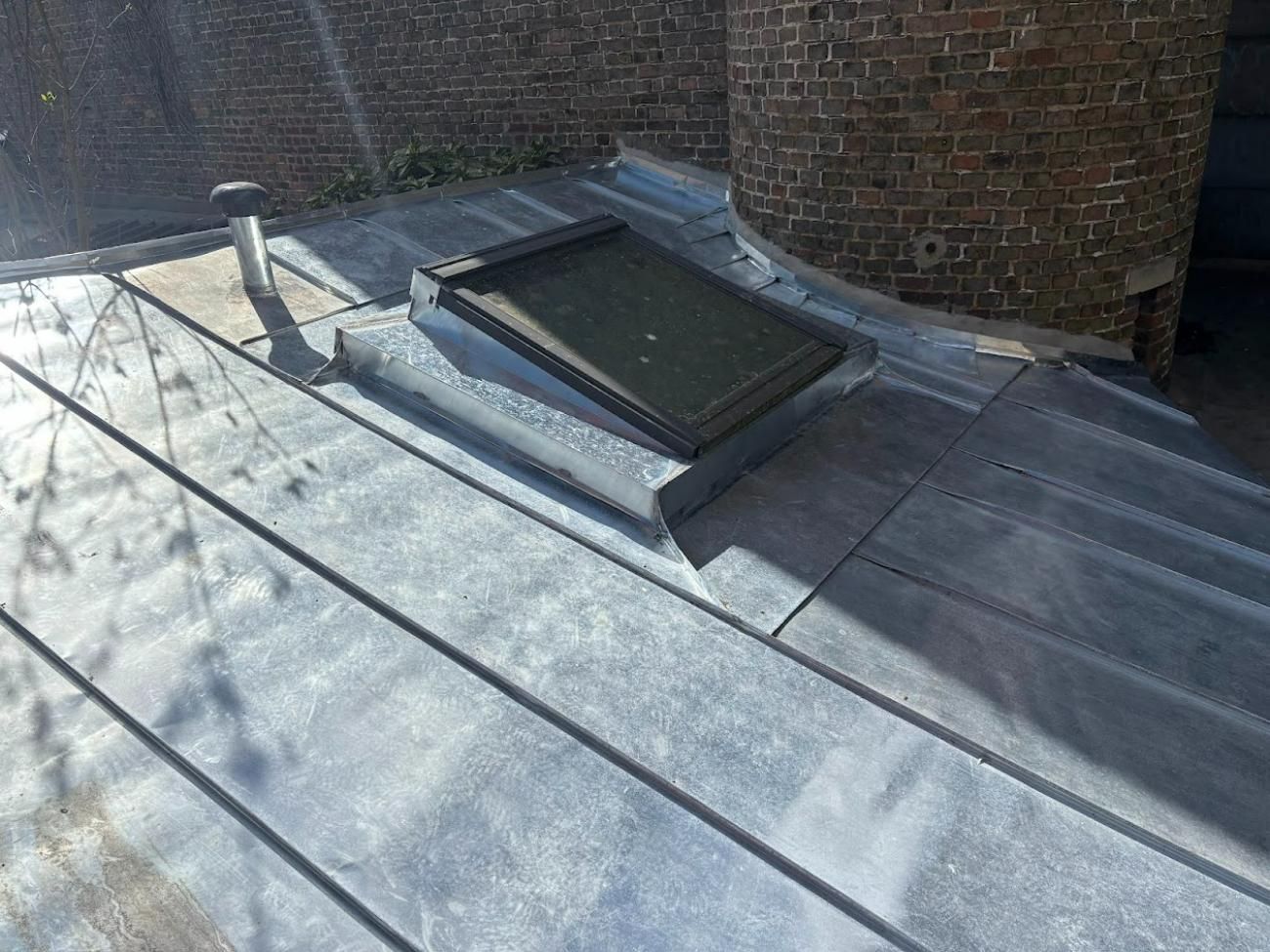 Un toit recouvert de plomb avec une lucarne et un mur de briques en arrière-plan, à l'extérieur.