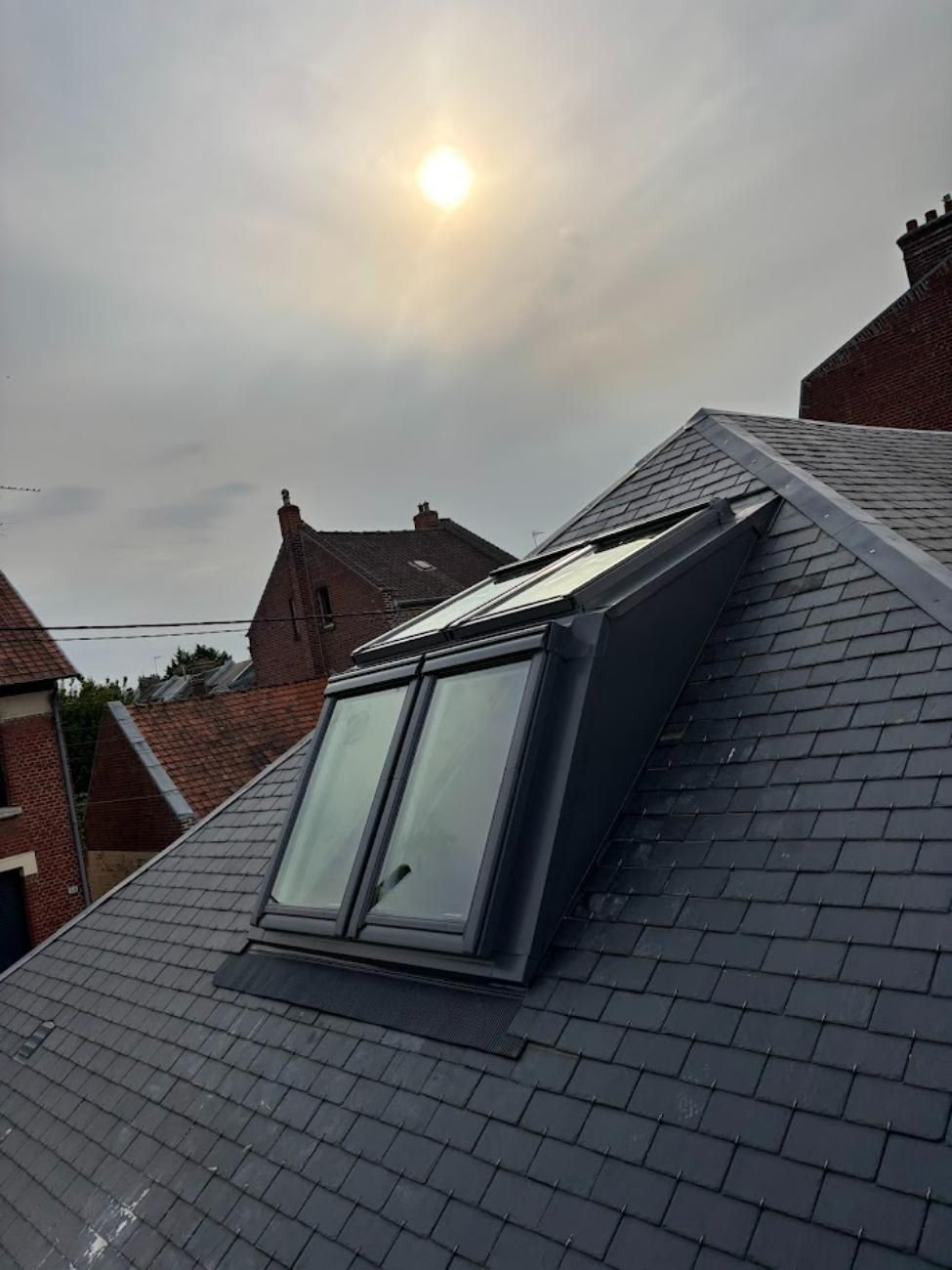 Puits de lumière sur un toit en ardoise, journée nuageuse, bâtiments en briques en arrière-plan, soleil visible.