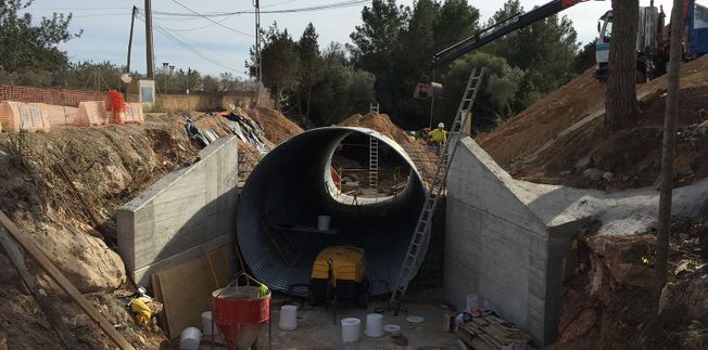 Sitio de construcción con un gran túnel cilíndrico en instalación; paredes de hormigón, grúa y trabajadores presentes.
