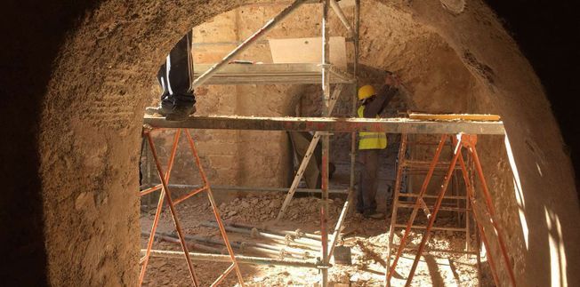 Trabajadores en un ascensor reparando un largo muro de piedra. Vista exterior con gente y maquinaria naranja.