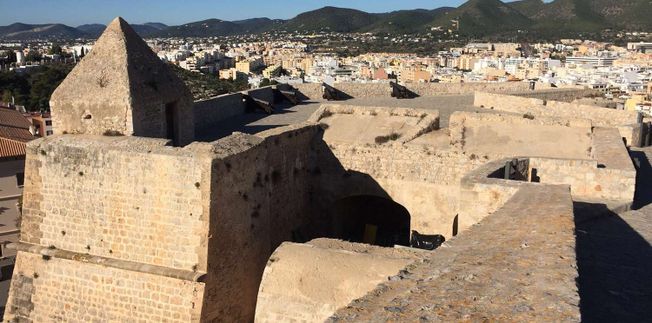 Fortaleza de piedra con vistas a una ciudad, montañas al fondo.