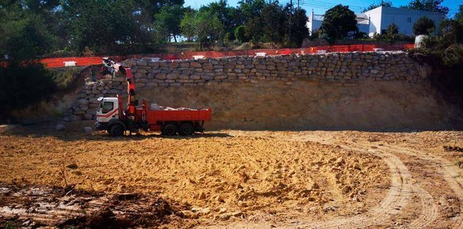 Una obra en construcción con un camión naranja descargando piedras en un muro de piedra. Campo de tierra.