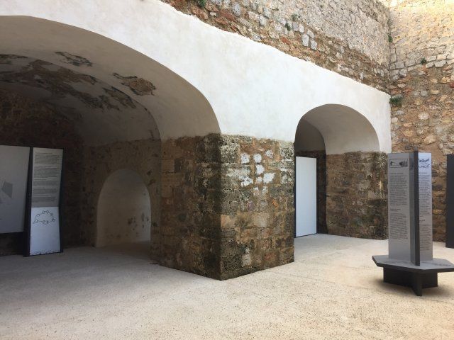 Interior abovedado con paredes de piedra, probablemente un museo. Techo blanco, exposiciones y un banco moderno.