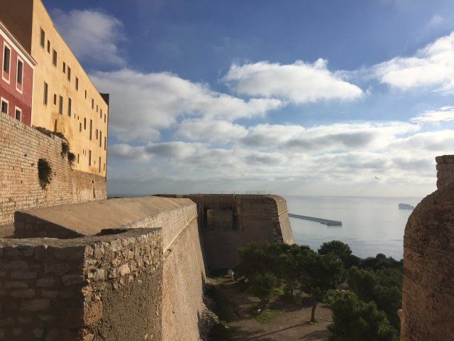 Muro de piedra y edificios con vistas al mar bajo un cielo azul nublado.