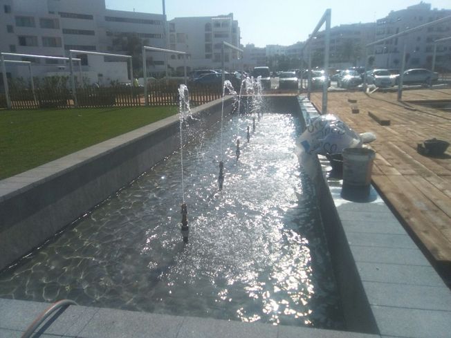 Una fuente de agua rectangular con chorros que rocían agua en un área pública con edificios al fondo.