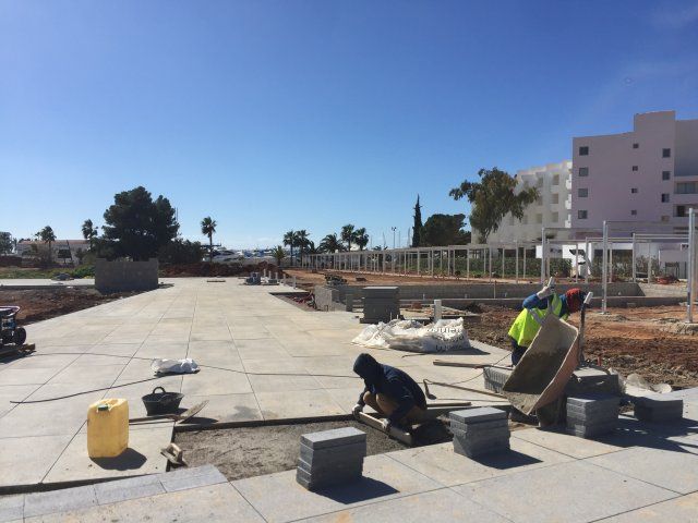 Obreros de la construcción colocando adoquines en un área abierta con un cielo azul y un edificio blanco al fondo.