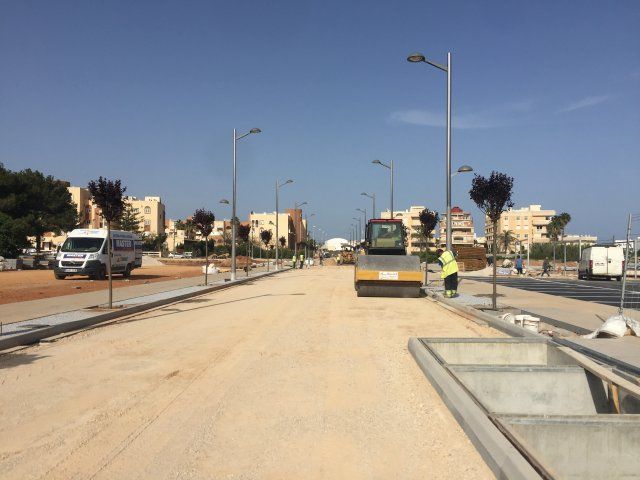 Construcción de carreteras; una apisonadora compacta grava. Se ven trabajadores y edificios. Día soleado.