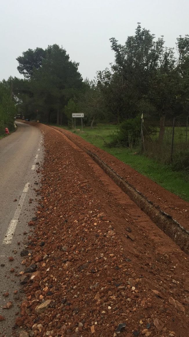 Construcción de la carretera: Tierra marrón cubre el arcén. Una zanja corre a lo largo del borde con una zona de césped y árboles al fondo.